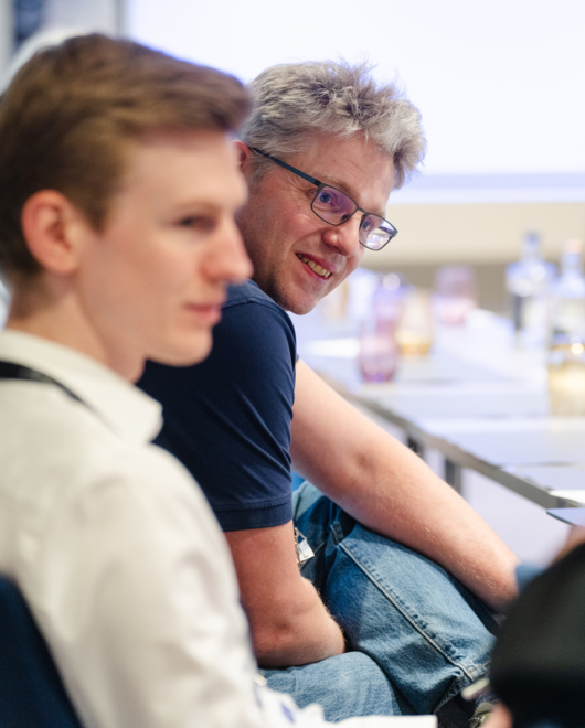 Zwei Männer in einem Meetingraum, einer mit kurzem, blonden Haar und einer mit grauem Haar, die sich freundlich unterhalten und lächeln.