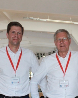 Zwei Männer in weißen Hemden mit roten Lanyards stehen in einem modernen Büro, lächeln und posieren. Der Raum hat große Fenster und eine offene Atmosphäre.